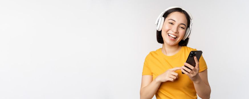 Cute japanese girl in headphones, looking at mobile phone and smiling, using music app on smartphone, standing against white background.