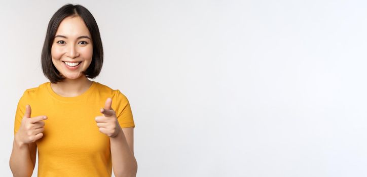 Young korean girl student, pointing fingers at camera and smiling, congratulating you, choosing, inviting people, standing over white background.