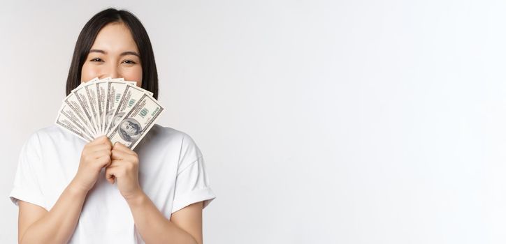 Portrait of smiling asian woman holding dollars money, concept of microcredit, finance and cash, standing over white background.