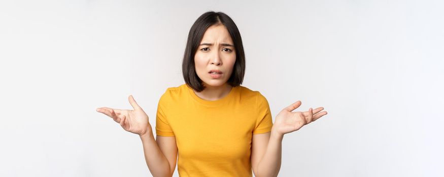 Frustrated, confused asian woman cant understand smth, looking clueless, shrugging shoulders, standing in yellow tshirt over white background.