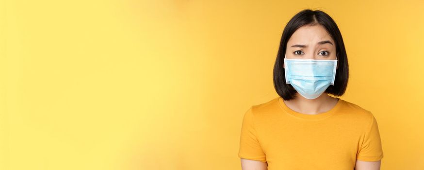 Portrait of skeptical and confused asian woman in medical face mask, raising eyebrow doubtful, standing over yellow background.