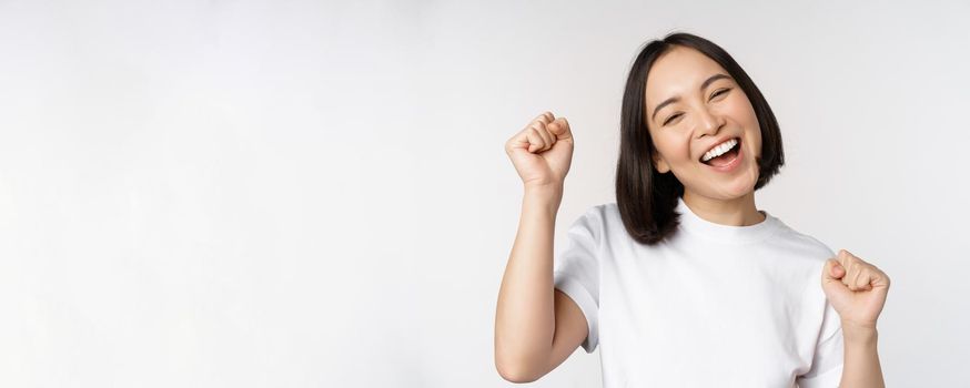 Dancing asian girl celebrating, feeling happy and upbeat, smiling broadly, standing over studio white background.