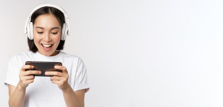 Happy asian woman in headphones, looking at smartphone, watching video on mobile phone and smiling, standing over white background.