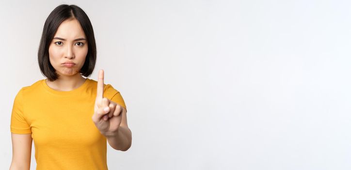 Portrait of asian woman looking serious and angry, showing stop prohibit gesture, taboo sign, forbidding smth, standing in yellow tshirt over white background.