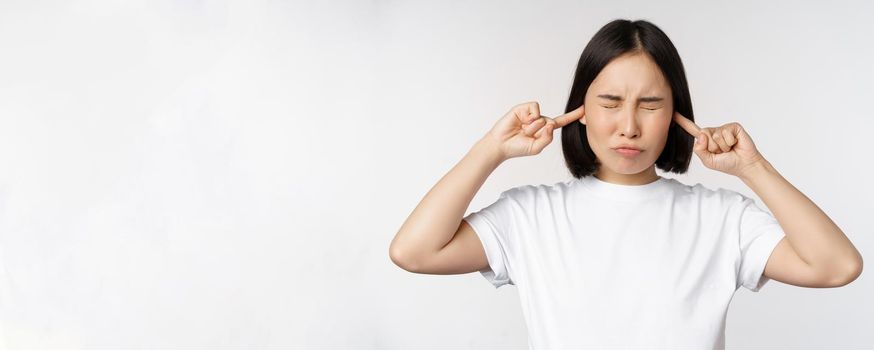 Portrait of asian woman shut ears and feeling discomfort from loud noise, annoying sound, standing over white background.