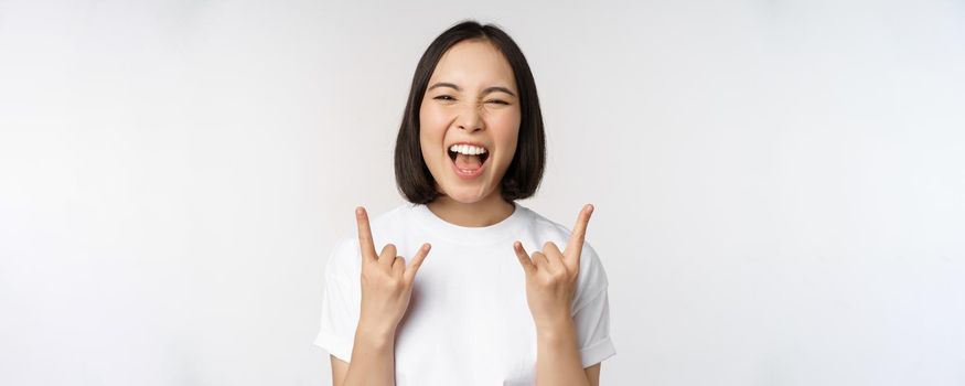 Sassy asian girl shouting, enjoying concert or festival, showing rock on, heavy metal sign, having fun, standing over white background.