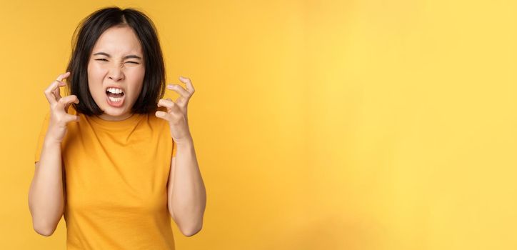 Image of angry asian woman, shouting and cursing, looking outraged, furious face expression, standing over yellow background.