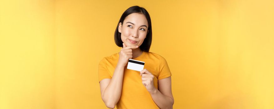 Thinking asian woman holding credit card and smiling, planning buy smth, standing thoughtful over yellow background.