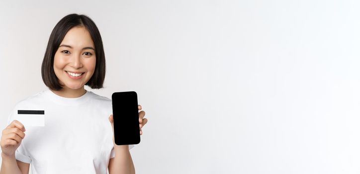 Portrait of smiling young asian woman showing mobile phone screen and credit card, standing over white background.