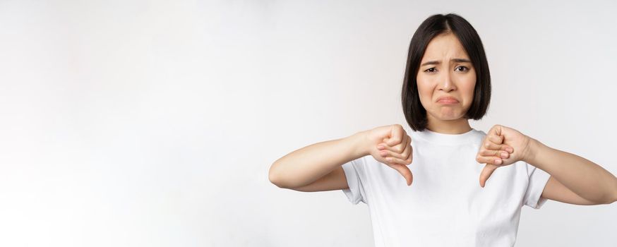 Image of asian woman showing thumbs down, dislike smth, looking disappointed, standing over white background. Copy space