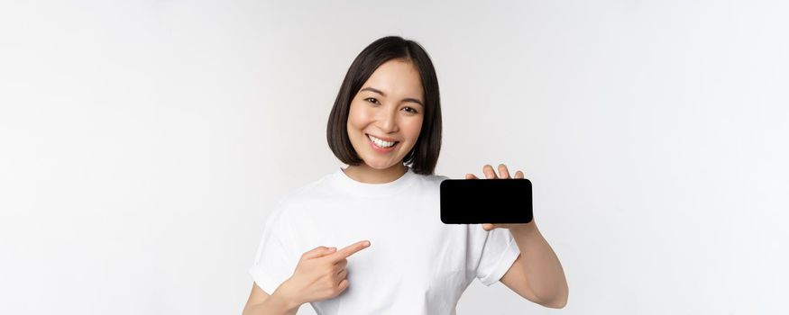 Portrait of smiling korean woman pointing finger at mobile phone screen, showing horizontal smartphone display, recommending website or store online, white background.