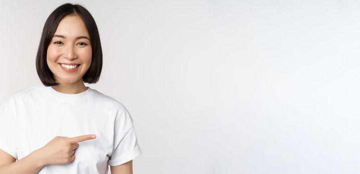 Happy beautiful asian girl showing advertisement on empty copy space, pointing finger right and smiling, demonstrating banner or company logo, white background.