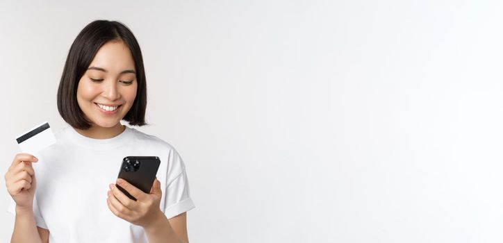 Online shopping. Happy asian woman using credit card and smartphone app, paying on website via mobile phone, white background.