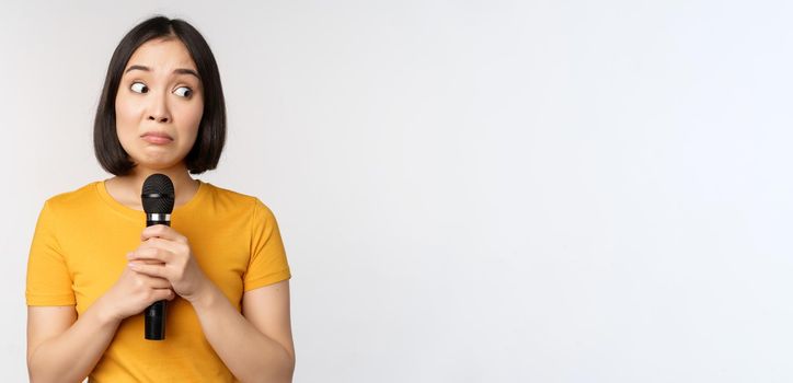 Modest asian girl holding microphone, scared talking in public, standing against white background. Copy space