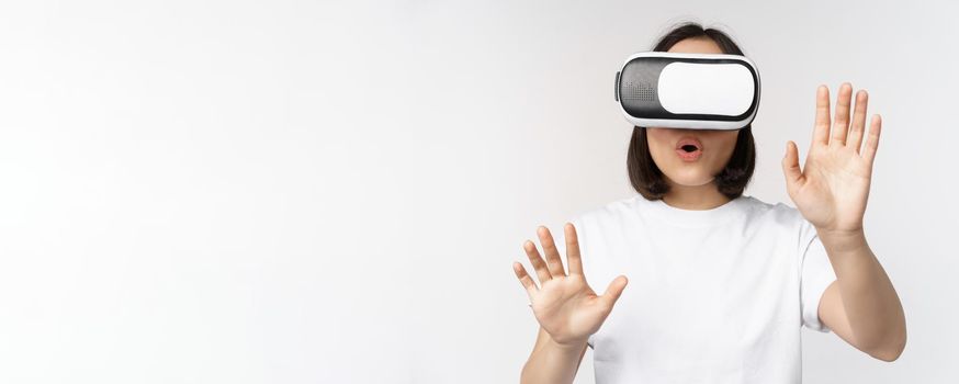 Amused asian girl using VR glasses, virtual reality headset and reaching hands into empty space, touching smth augmented, standing over white background.