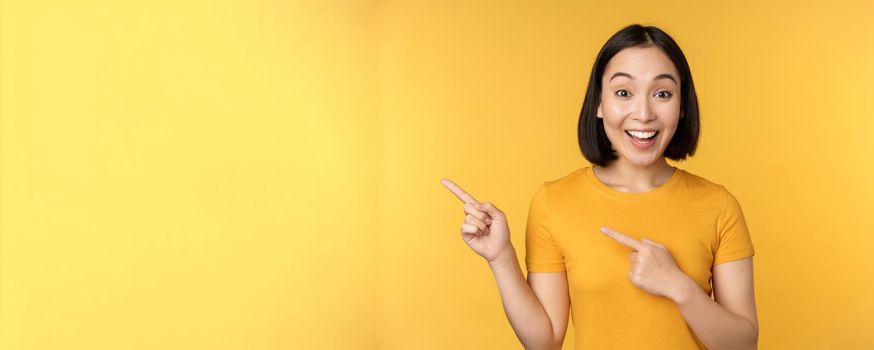 Smiling asian woman pointing fingers left, showing advertisement on empty copy space, standing over yellow background.