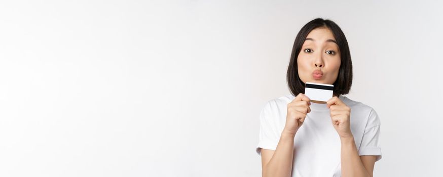 Money and finance concept. Cute japanese girl kissing her credit card, standing in tshirt over white background.