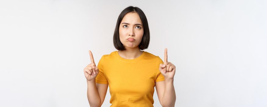 Disappointed asian woman looking, pointing fingers up with angry moody face expression, standing in yellow tshirt over white background.