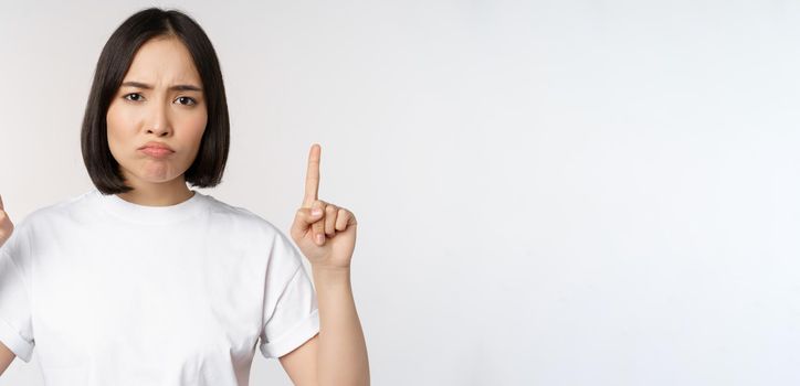 Sad and disappointed young asian woman pointing fingers up, showing advertisement with upset face expression, white studio background.
