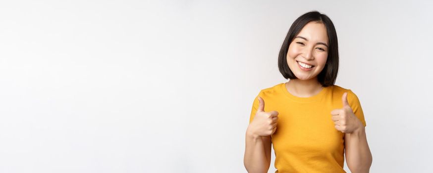 Beautiful smiling asian female model, showing thumbs up and looking pleased, recommending, express positive feedback, standing over white background.