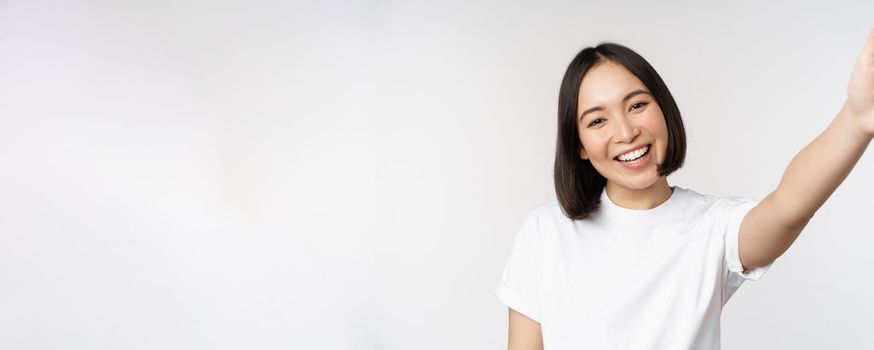 Beautiful young asian woman smiling, looking at camera, holding device, taking selfie, video chat, standing in tshirt over white background.