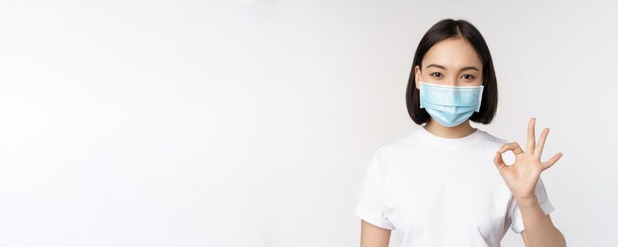 Covid-19, healthcare and medical concept. Happy asian girl smiling, wearing medical face mask, showing okay, ok sign, approve, recommending smth, white background.