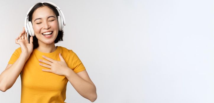 Happy asian girl dancing, listening music on headphones and smiling, standing in yellow tshirt against white background. Copy space