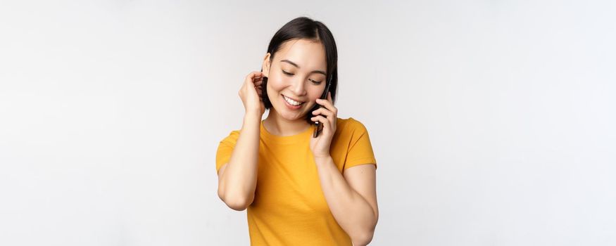 Romantic and cute asian girl answer phone call, talking on mobile, smiling coquettish, standing in yellow tshirt against white background.