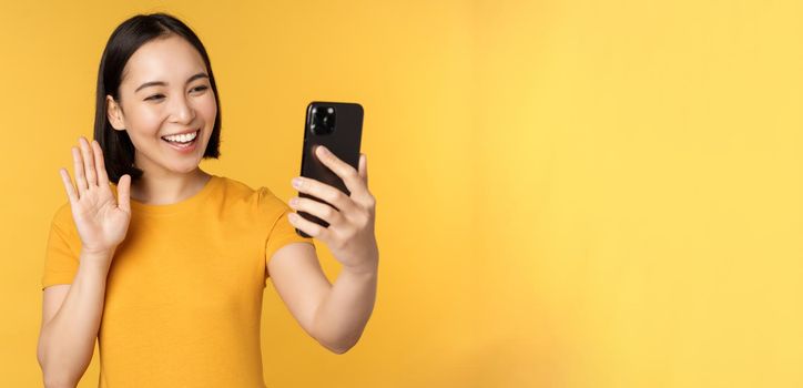 Image of happy, beautiful asian girl video chatting, talking on smartphone application, standing against yellow background. Copy space