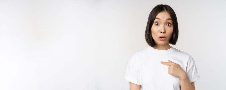 Portrait of surprised asian girl student, pointing finger left, raising eyebrows amazed, impressed by big deal, offer ahead, standing over white background.