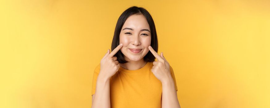 Close up portrait of cute asian girl showing her dimples and smiling coquettish at camera, standing over yellow background. Copy space
