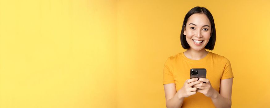 Technology. Smiling asian woman using mobile phone, holding smartphone in hands, standing in t-shirt against yellow background.