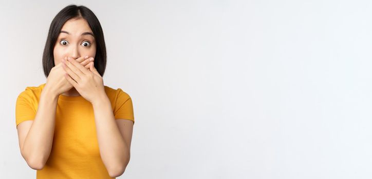 Shocked asian woman cover mouth with hands, looking startled with speechless face expression, standing in yellow tshirt against white background.