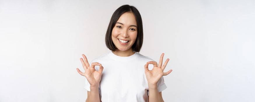Very well, excellent. Smiling asian woman showing okay sign, approval, ok gesture, looking satisfied, recommending smth, standing over white background.