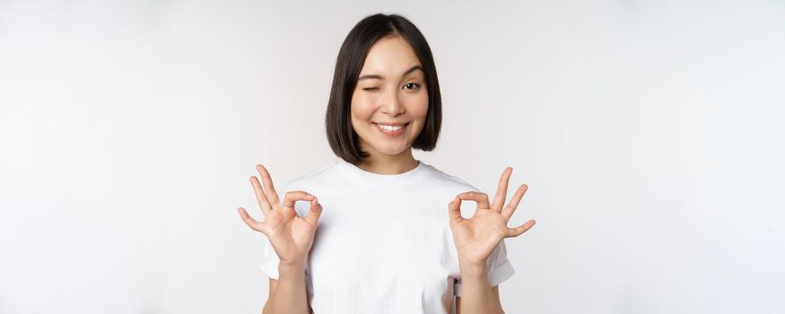 Very well, excellent. Smiling asian woman showing okay sign, approval, ok gesture, looking satisfied, recommending smth, standing over white background.