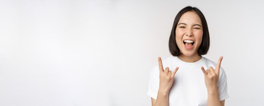 Sassy asian girl shouting, enjoying concert or festival, showing rock on, heavy metal sign, having fun, standing over white background.