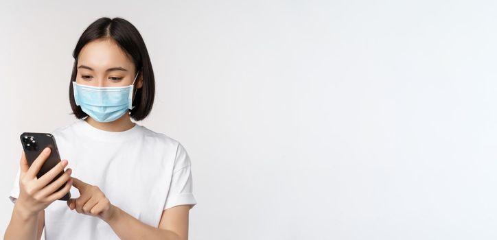 Health and covid-19 concept. Young asian woman in medical mask using mobile phone, typing on smartphone, standing against white background.