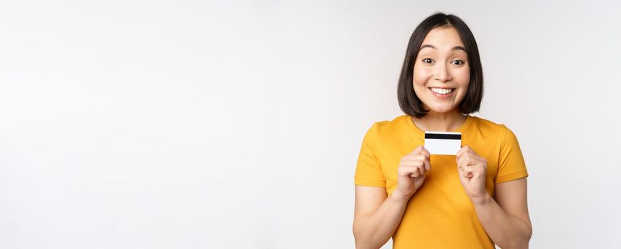 Portrait of beautiful korean girl holding credit card, recommending bank service, standing in yellow tshirt over white background.