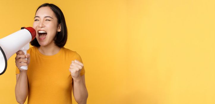 Announcement. Happy asian woman shouting loud at megaphone, recruiting, protesting with speaker in hands, standing over yellow background.