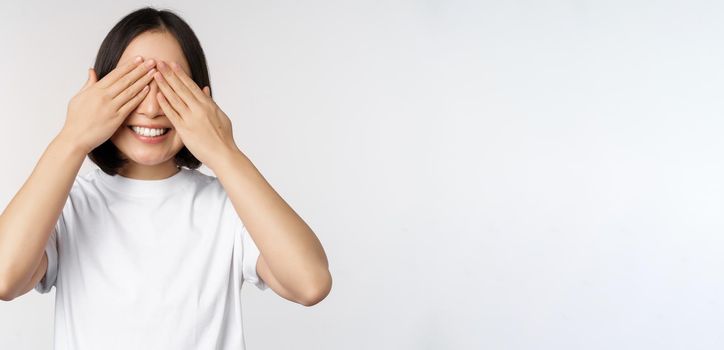 Portrait of asian woman covering eyes, waiting for surprise blindfolded, smiling happy, anticipating, standing against white background.