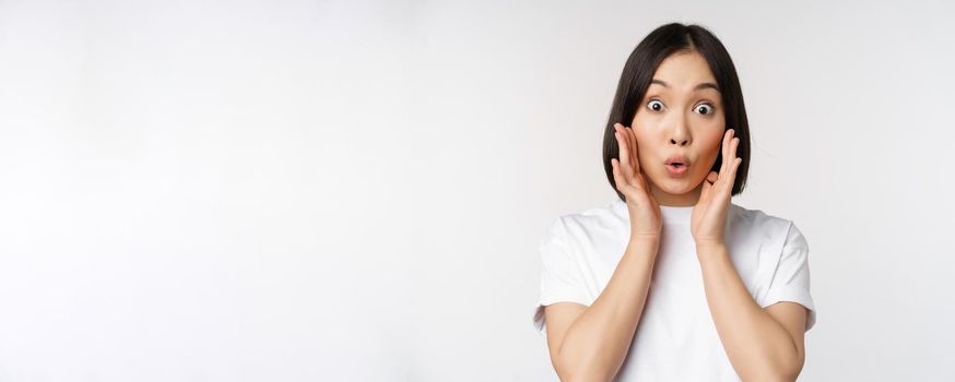 Close up portrait of asian girl looking surprised, wow face, reacting amazed at smth, standing in white tshirt over studio background, isolated.