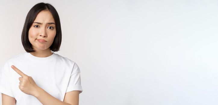 Skeptical asian girl in white t-shirt, pointing at product or logo with disappointed grimace, dislike and complain at smth, standing over white background.