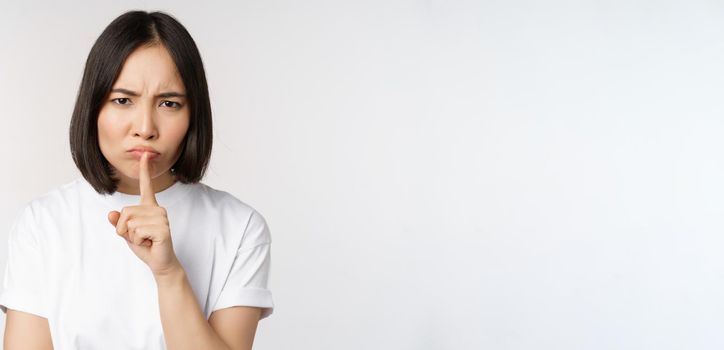 Angry asian girl shushing, keep quiet, taboo silence gesture, press finger to lips and frowning, scolding for being too loud, standing over white background.