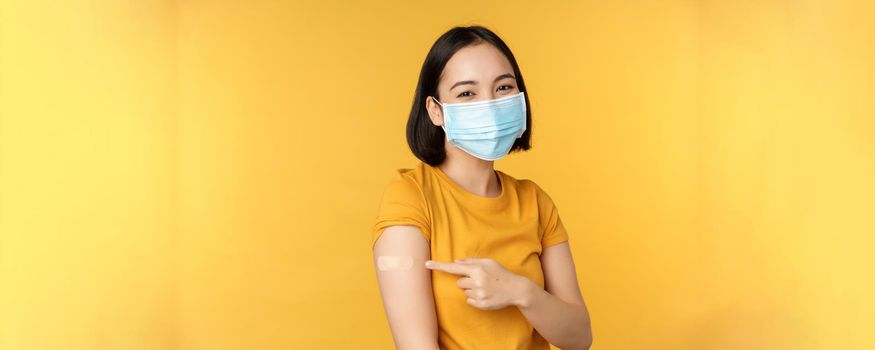 Vaccination and covid-19 pandemic concept. Smiling asian woman in medical face mask, showing her shoulder with band aid after vaccinating from coronavirus, yellow background.