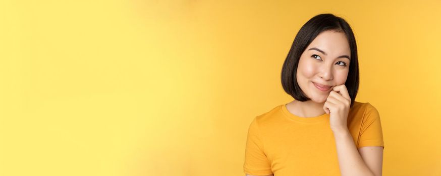 Close up portrait of cute asian girl smiling, thinking, looking up thoughtful, standing in tshirt over yellow background. Copy space