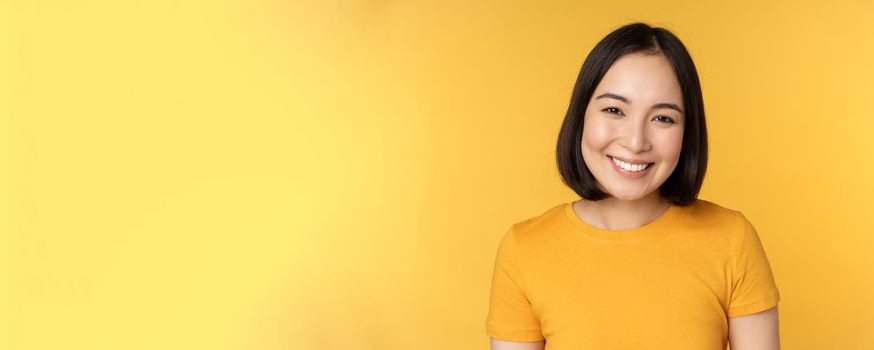 Close up portrait of beautiful asian woman smiling, looking cute and tender, standing against yellow background.