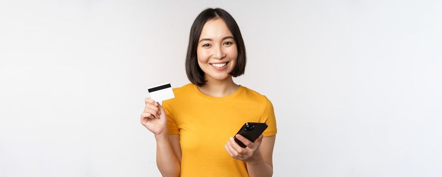 Beautiful smiling asian girl using credit card and mobile phone, paying online on smartphone, standing in yellow tshirt over white background.