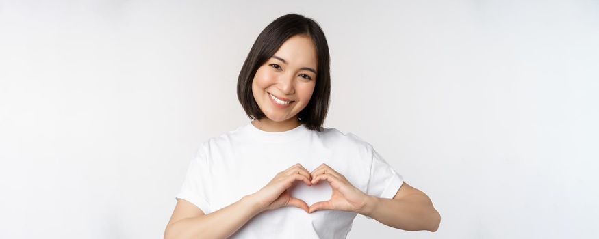 Lovely asian woman smiling, showing heart sign, express tenderness and affection, standing over white background.