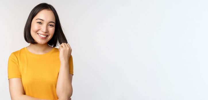 Beautiful romantic asian girl, smiling and playing with hair, looking happy at camera, standing in yellow t-shirt over white background.