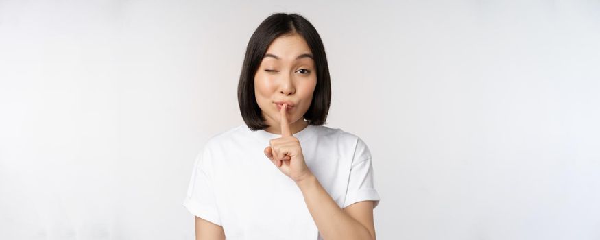Close up portrait of young beautiful asian girl shushing, has secret, keep quiet silence gesture, press finger to lips, standing in tshirt over white background.